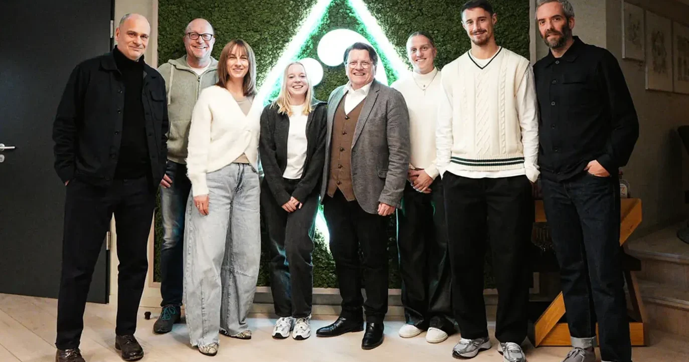Eine Gruppe von sieben Personen steht vor einem grünen Hintergrund mit einem dreieckigen Muster. Die Personen lächeln und posieren für ein Gruppenfoto. Sie tragen verschiedene Outfits, darunter Jeans, Pullover und Blazer. Der Raum wirkt modern und hell.