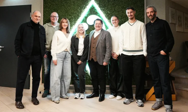 Eine Gruppe von sieben Personen steht vor einem grünen Hintergrund mit einem dreieckigen Muster. Die Personen lächeln und posieren für ein Gruppenfoto. Sie tragen verschiedene Outfits, darunter Jeans, Pullover und Blazer. Der Raum wirkt modern und hell.