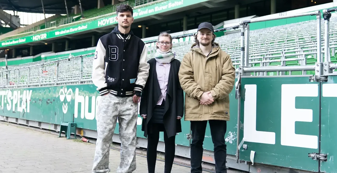Drei Personen stehen vor einer grünen Stadionwand mit der Aufschrift "Hoyer" und "LE". Die Person links trägt ein schwarzes Jackett mit einem großen "B" auf der Brust, die Person in der Mitte trägt einen schwarzen Mantel mit einem Schal und die Person rechts trägt eine beige Jacke und eine schwarze Mütze. Im Hintergrund sind grüne Sitzreihen des Stadions zu sehen.