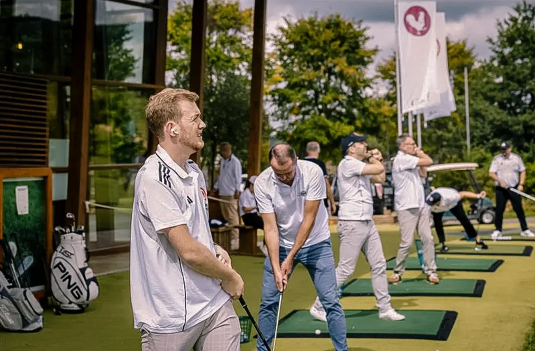 Eine Gruppe von Männern spielt Golf auf einer Indoor-Golfanlage. Sie stehen auf grünen Teppichen und üben ihre Schläge. Im Hintergrund sind weitere Personen zu sehen, darunter eine mit einem Golfbag. Eine Flagge mit einem Herz-Logo hängt an der Wand.