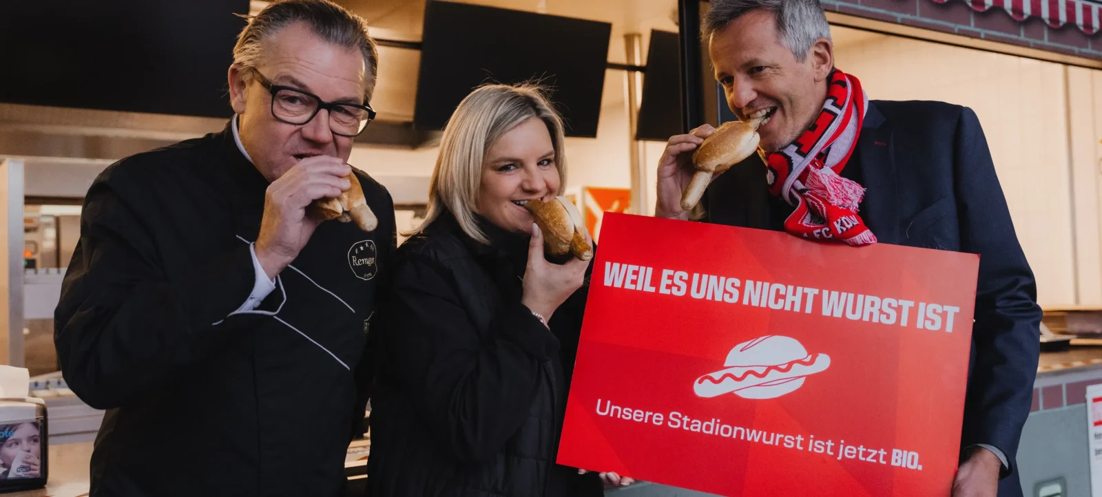 Drei Personen essen Wurstbrötchen und halten ein Schild mit der Aufschrift "WEIL ES UNS NICHT WURST IST. Unsere Stadionwurst ist jetzt BIO.