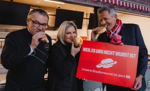 Drei Personen essen Wurstbrötchen und halten ein Schild mit der Aufschrift "WEIL ES UNS NICHT WURST IST. Unsere Stadionwurst ist jetzt BIO.