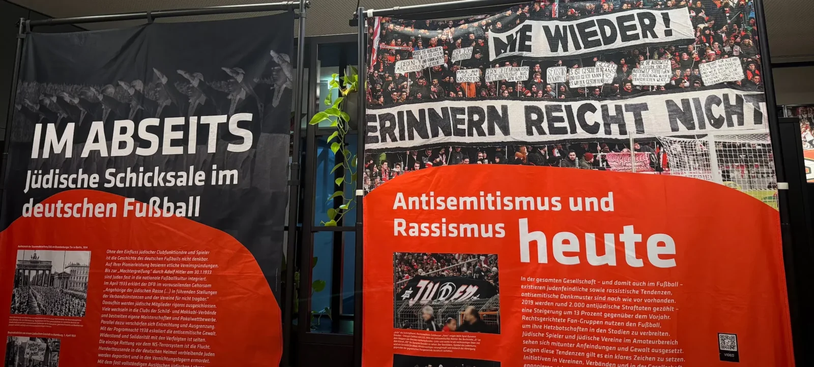 Ausstellung über Antisemitismus und Rassismus im Fußball. Links ein Plakat mit dem Titel 'Im Abseits: Jüdische Schicksale im deutschen Fußball' zeigt historische Fotos und Texte zur Rolle jüdischer Spieler und Funktionäre im deutschen Fußball während der NS-Zeit. Rechts ein Plakat mit dem Titel 'Antisemitismus und Rassismus heute' zeigt aktuelle Fotos und Texte zu antisemitischen und rassistischen Vorfällen in der Gegenwart, einschließlich einer Demonstration mit dem Spruch 'Wir erinnern: Grüß Gott nicht'.
