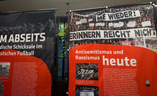 Ausstellung über Antisemitismus und Rassismus im Fußball. Links ein Plakat mit dem Titel 'Im Abseits: Jüdische Schicksale im deutschen Fußball' zeigt historische Fotos und Texte zur Rolle jüdischer Spieler und Funktionäre im deutschen Fußball während der NS-Zeit. Rechts ein Plakat mit dem Titel 'Antisemitismus und Rassismus heute' zeigt aktuelle Fotos und Texte zu antisemitischen und rassistischen Vorfällen in der Gegenwart, einschließlich einer Demonstration mit dem Spruch 'Wir erinnern: Grüß Gott nicht'.
