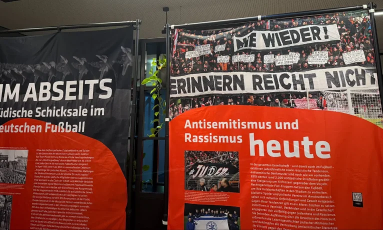 Ausstellung über Antisemitismus und Rassismus im Fußball. Links ein Plakat mit dem Titel 'Im Abseits: Jüdische Schicksale im deutschen Fußball' zeigt historische Fotos und Texte zur Rolle jüdischer Spieler und Funktionäre im deutschen Fußball während der NS-Zeit. Rechts ein Plakat mit dem Titel 'Antisemitismus und Rassismus heute' zeigt aktuelle Fotos und Texte zu antisemitischen und rassistischen Vorfällen in der Gegenwart, einschließlich einer Demonstration mit dem Spruch 'Wir erinnern: Grüß Gott nicht'.