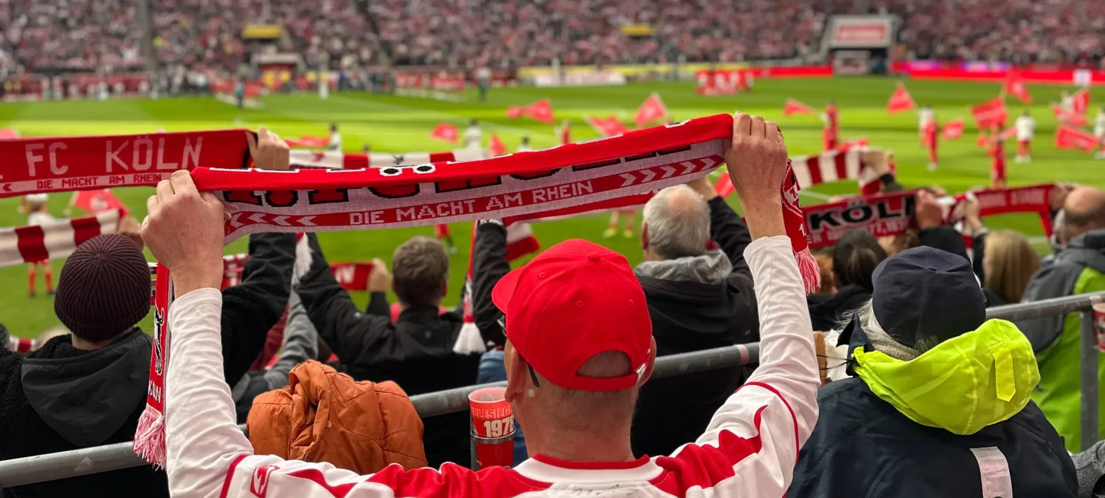 Ein Fan des FC Köln hält ein rotes Schal mit dem Aufdruck "FC Köln - Die Macht am Rhein" hoch. Im Hintergrund ist das Stadion mit vielen Zuschauern und dem Spielfeld zu sehen.