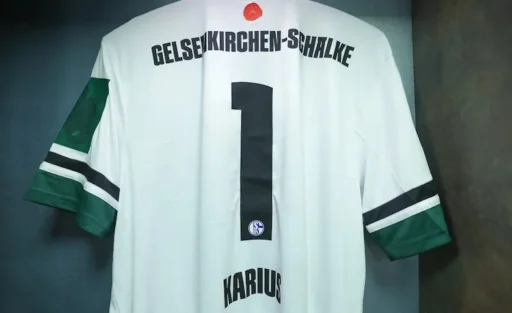 Ein weißes Trikot des Fußballvereins FC Schalke 04 mit grünen Ärmeln und der Nummer 1 auf dem Rücken. Der Name "Karius" ist unter der Nummer zu sehen. Das Trikot hat einen schwarzen Streifen auf dem Rücken und einen roten Knopf am Kragen.