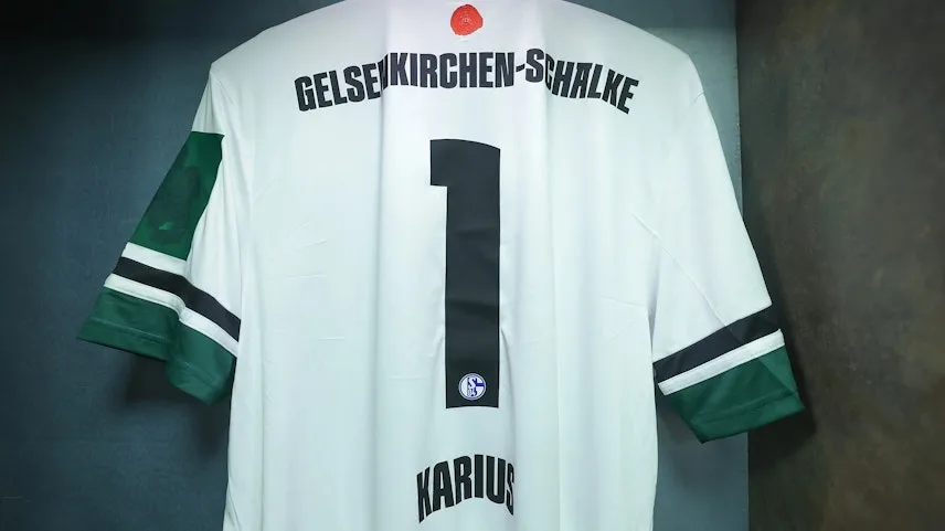 Ein weißes Trikot des Fußballvereins FC Schalke 04 mit grünen Ärmeln und der Nummer 1 auf dem Rücken. Der Name "Karius" ist unter der Nummer zu sehen. Das Trikot hat einen schwarzen Streifen auf dem Rücken und einen roten Knopf am Kragen.