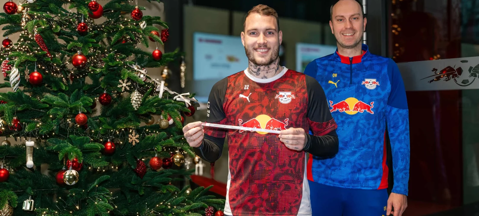 Zwei Personen in Trikots des RB Leipzig vor einem geschmückten Weihnachtsbaum. Die Person links hält ein weißes Band in den Händen.