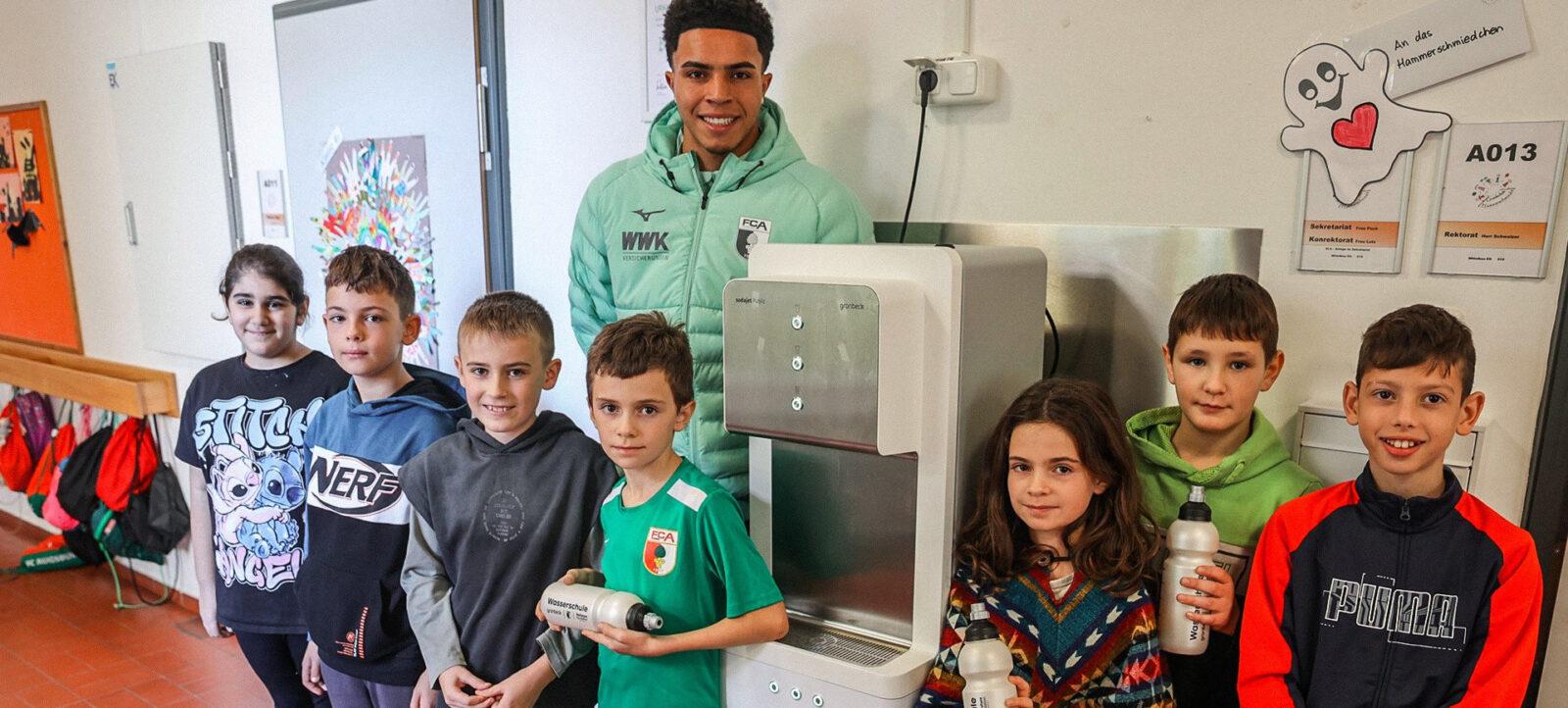 Ein junger Fußballspieler in einem grünen Trainingsanzug steht mit sieben Kindern vor einem Trinkbrunnen in einem Schulflur. Die Kinder halten Trinkflaschen in den Händen und lächeln in die Kamera. Im Hintergrund sind bunte Bilder und ein Schild mit einem herzlichen Symbol zu sehen.