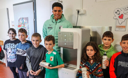 Ein junger Fußballspieler in einem grünen Trainingsanzug steht mit sieben Kindern vor einem Trinkbrunnen in einem Schulflur. Die Kinder halten Trinkflaschen in den Händen und lächeln in die Kamera. Im Hintergrund sind bunte Bilder und ein Schild mit einem herzlichen Symbol zu sehen.